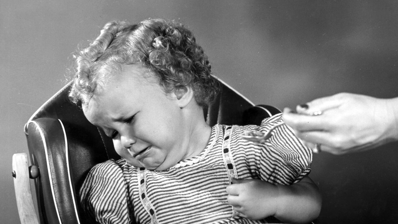 A baby girl sitting in a high chair turning her head away from a spoonful of food.