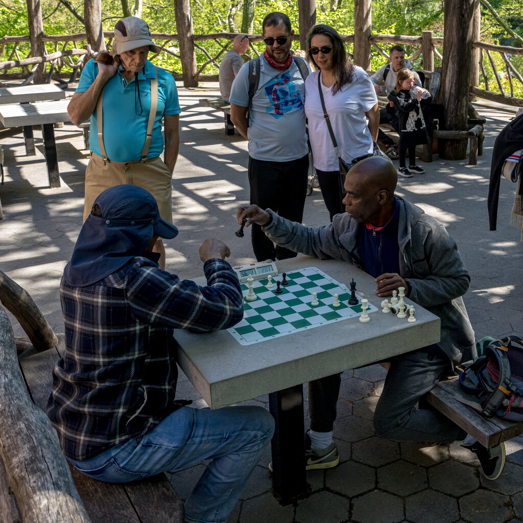 Three people stand around a table watching two men play chess.