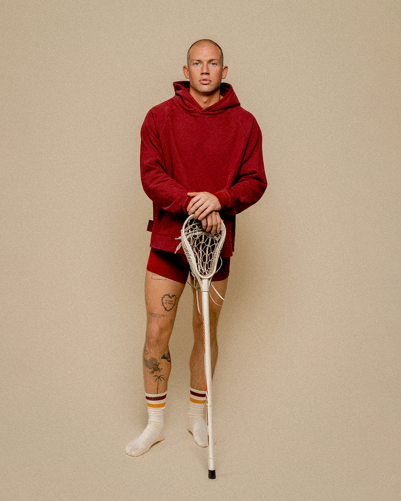 Man in studio wearing burgundy hoodie and burgundy boxer briefs holding field hockey stick