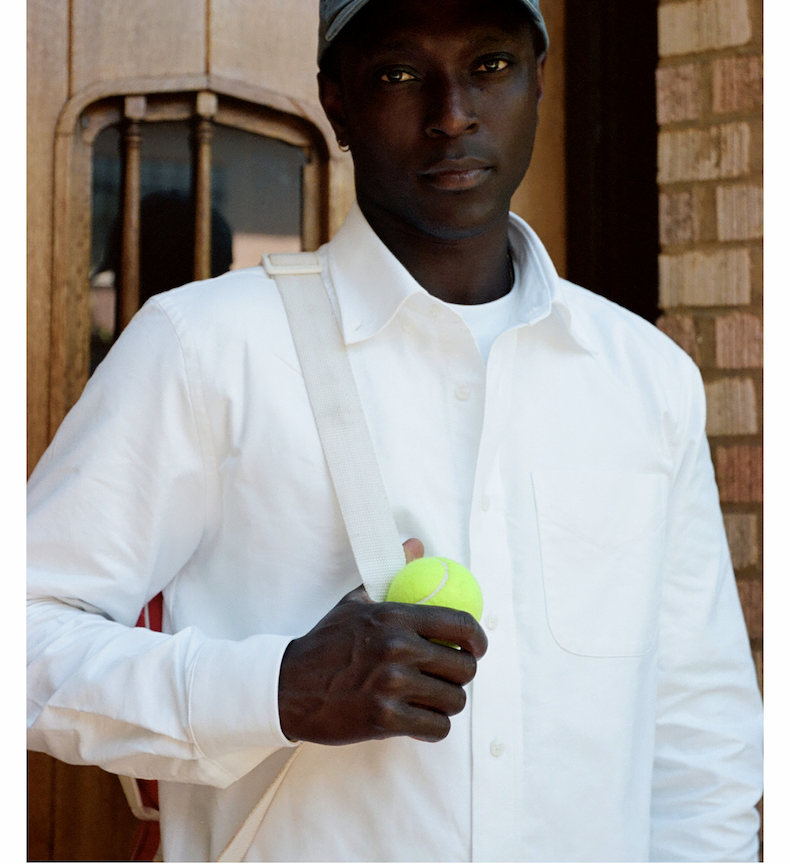 Man wearing a white oxford shirt and gray cap, holding a tennis ball with a bag strap over his shoulder.