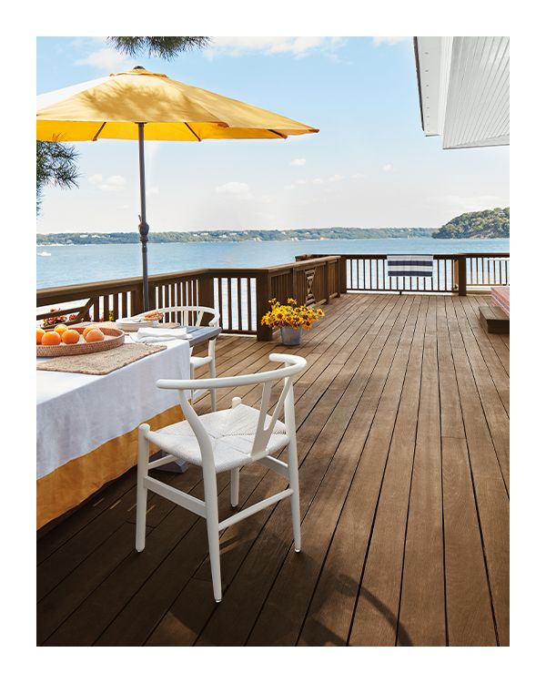 A beautiful waterside brown stained deck with a dining table, white painted chairs and a yellow umbrella.
