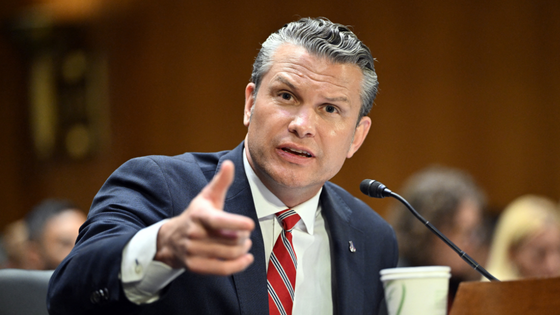 A man speaks into a microphone and points at the camera.