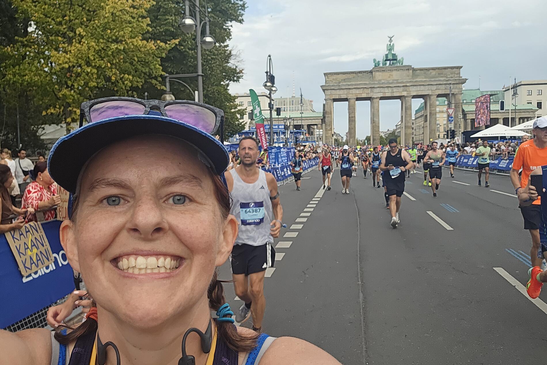 Lydia Schrandt smiling as she takes a selfie while running a race in a city amid other runners wearing race bibs. Onlookers cheer racers on from the sidelines.