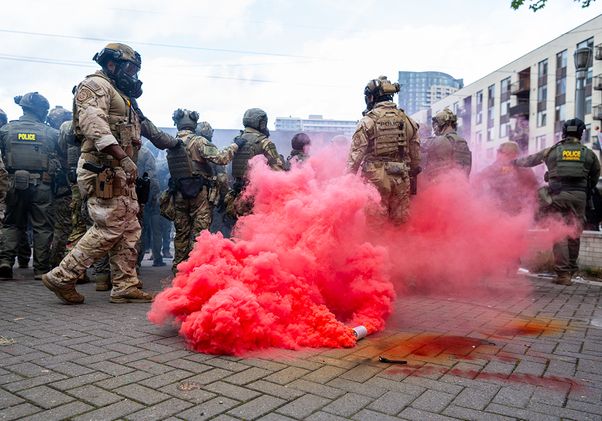 Federal officers wearing gas masks standing near red smoke billowing from a tear gas canister