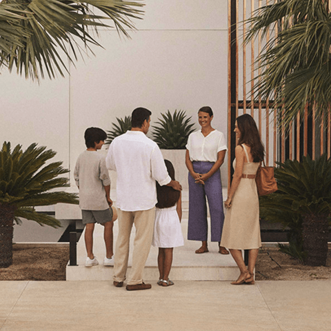 Residence staff member greeting a family at the entrance steps, surrounded by palm plants and white walls.