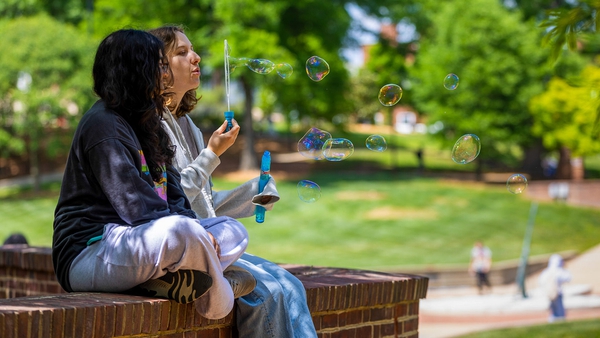 two students sit on brick ledge and blow bubbles