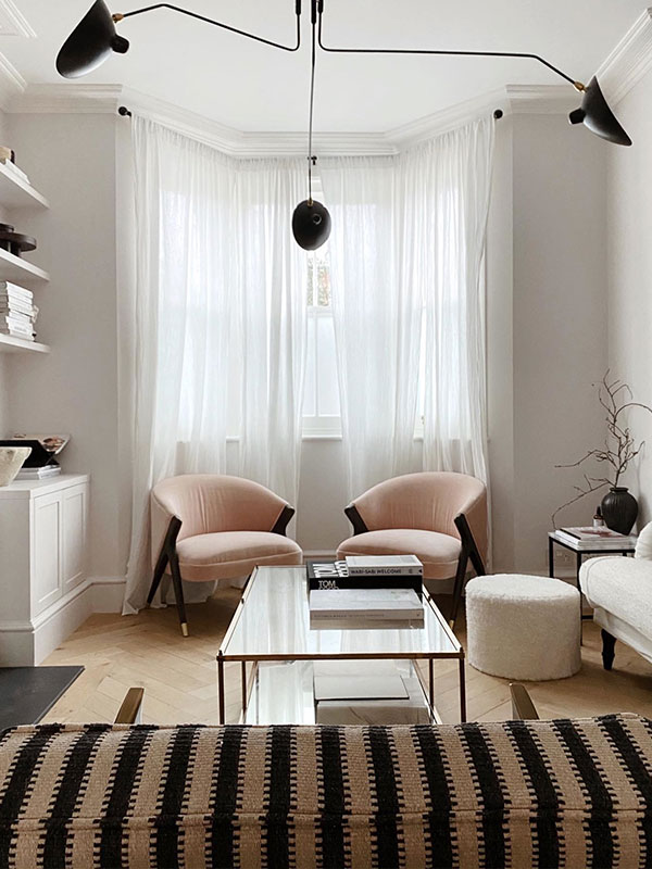A bright corner with two blush pink chairs, sheer white curtains, and a glass coffee table. Clean lines and soft tones give a refined, contemporary look.