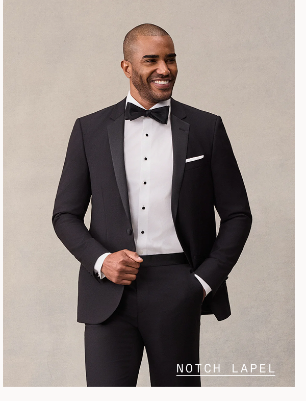 Man with a shaved head smiles, wearing a black tuxedo with satin notch lapels, black bow tie, white tuxedo shirt with black buttons, and white pocket square, hand in pocket. Text overlay reads: "NOTCH LAPEL"