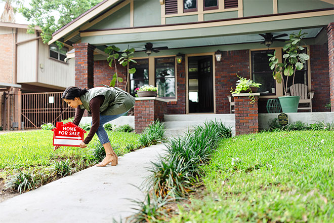 A person places a "Home - For Sale" sign on their front lawn.
