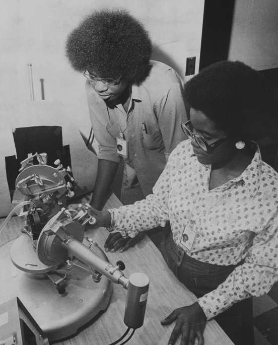 Greyscale photo of two Black students from the 1970s working with a small machine on a lab bench
