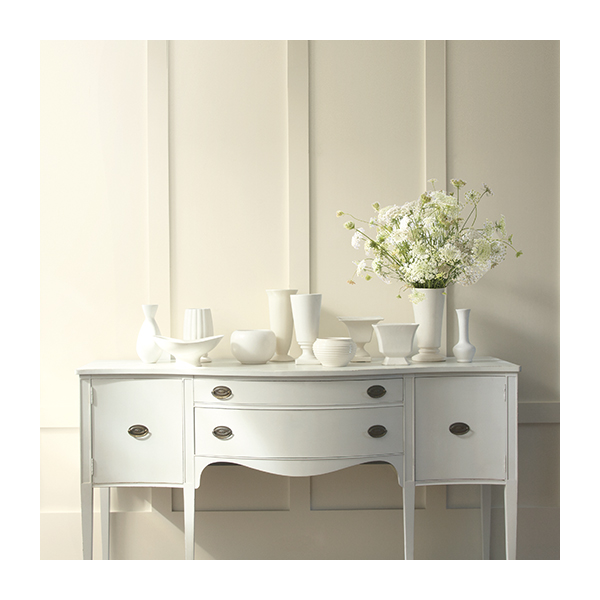An off-white wall with floor-to-ceiling wainscoting and multiple white vases placed on top of a white dresser.