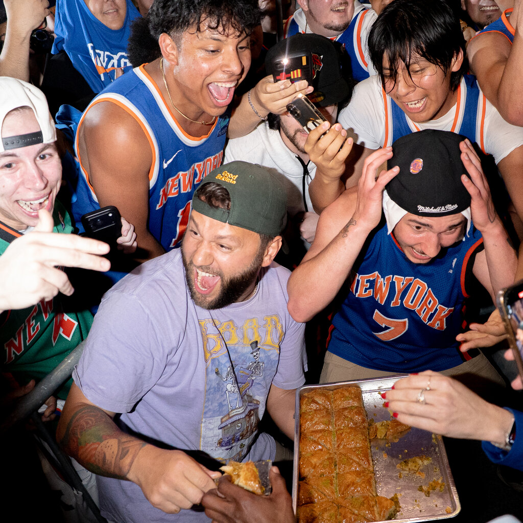 Emotional fans in Knicks jerseys surround the Baklava Guy, who is wearing a lavender T-shirt holding a tray of baklava.