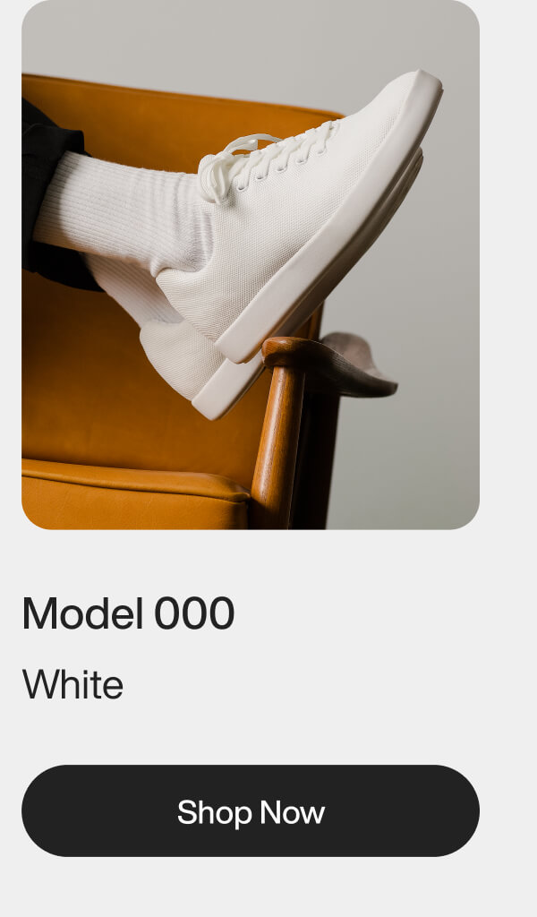 A white sneaker, worn with white socks, rests on an orange chair with wooden arms. Below are the words "Model 000 White" and a "Shop Now" button.