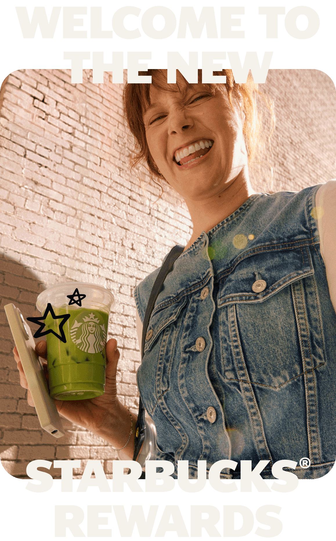 Red‑haired person in a denim vest holds a green iced Starbucks drink and a phone in front of a brick wall, with star doodles around the cup. Bold text reads “Welcome to the new Starbucks Rewards.”