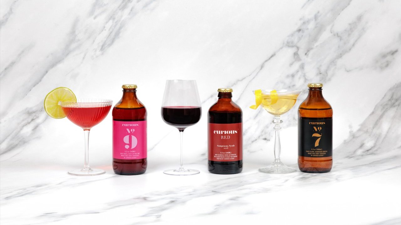 A bottle of Curious No. 9, Curious Red, and Curious No. 7 lined up next to it's served drink on a marble background.