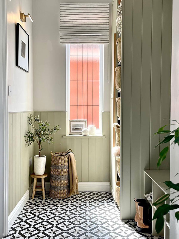A compact hallway featuring sage green panelling, black‑and‑white geometric floor tiles, and woven baskets. Natural light and plants add freshness.