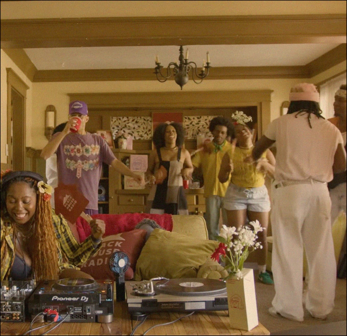  DJ 'Packrat' on a mixer and turntable setup in a vibrant home while people party in the background.