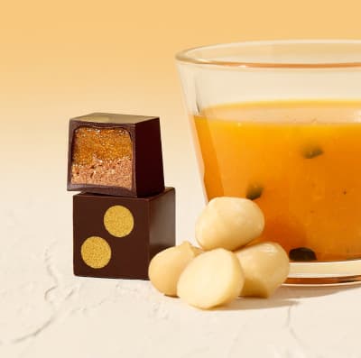 Two Passion Fruit & Macadamia bonbons next to a glass of passion fruit juice and a few macadamias.
