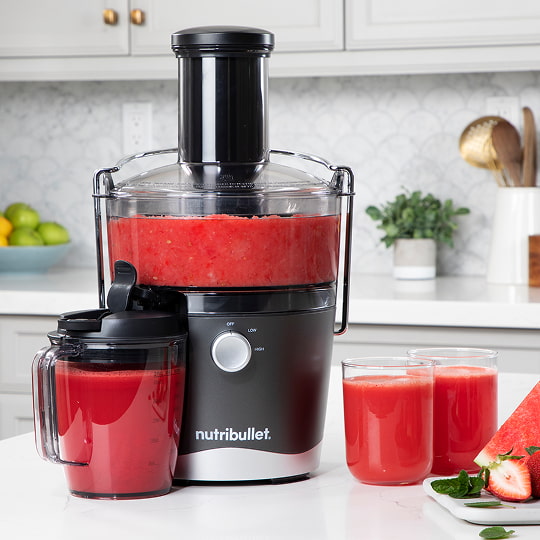  A nutribullet Juicer™ with red juice sits on a kitchen counter. Nearby are a pitcher, two glasses, and a cutting board with fruit.