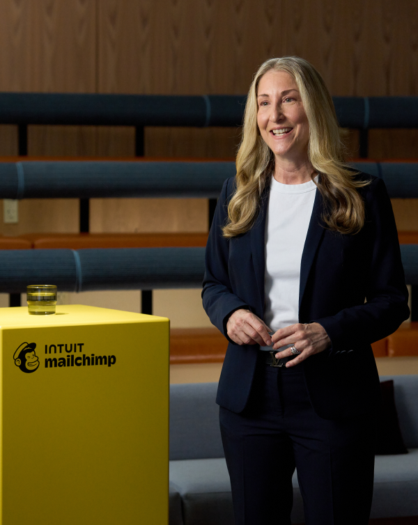 An icon of a megaphone, followed by the headline “Join the excitement.” Below, text reads “Unlock exclusive access to these fresh insights—all at no cost with your RSVP.” The time listed reads “12-1 pm UTC-4.” There is a call to action reading “Register now.”

To the right, speaker Tiffani Bova stands next to a yellow pedestal with the Intuit Mailchimp logo and a glass of water on it. She is wearing a navy blue blazer and a white shirt. 