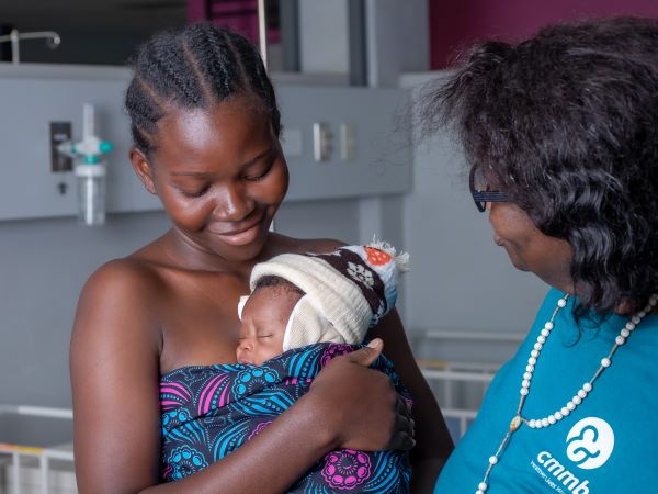 Alt-text: Memory, a mother in Zambia, hold her infant son under the supervision of a health care worker