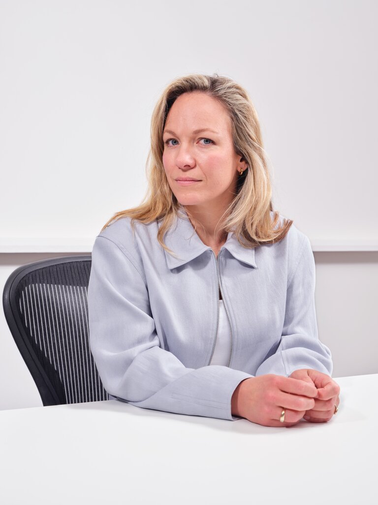 A woman in a blue jacket and white shirt, seated in an office chair behind a table, with her face turned but looking into the camera.