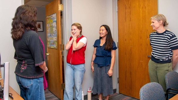 two students smile, with one holding hands to her face, as two other people look on