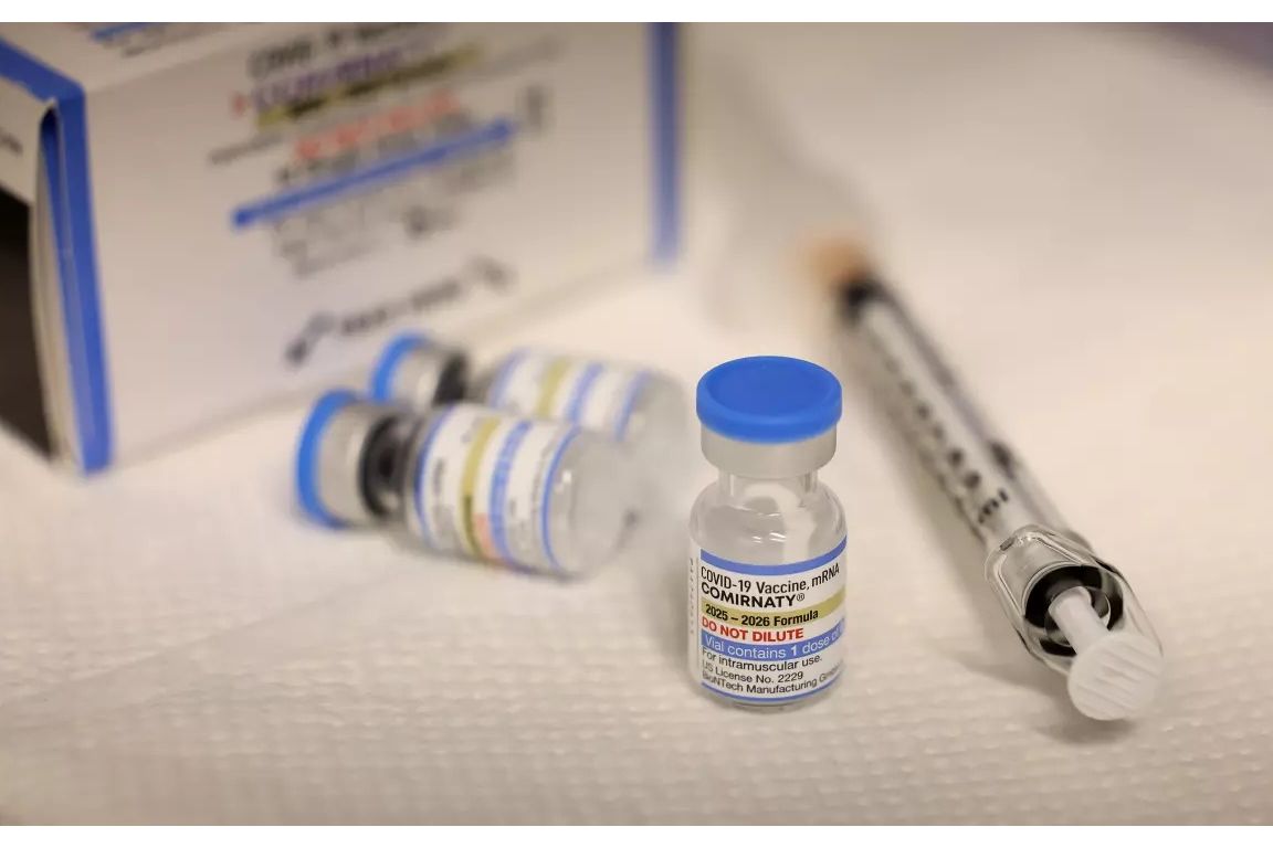 Close-up of a COVID-19 vaccine vial labeled Comirnaty beside a syringe, with additional vials and packaging blurred in the background