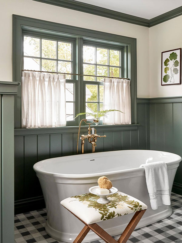 A freestanding white bathtub beneath green‑framed windows with short white curtains. Brass fixtures and leafy accents add a timeless, elegant touch.