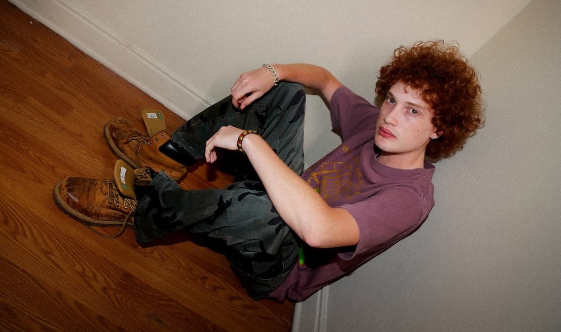 Model sitting against a beige wall wearing a muted purple Obey t-shirt, dark cargo pants, and work boots.