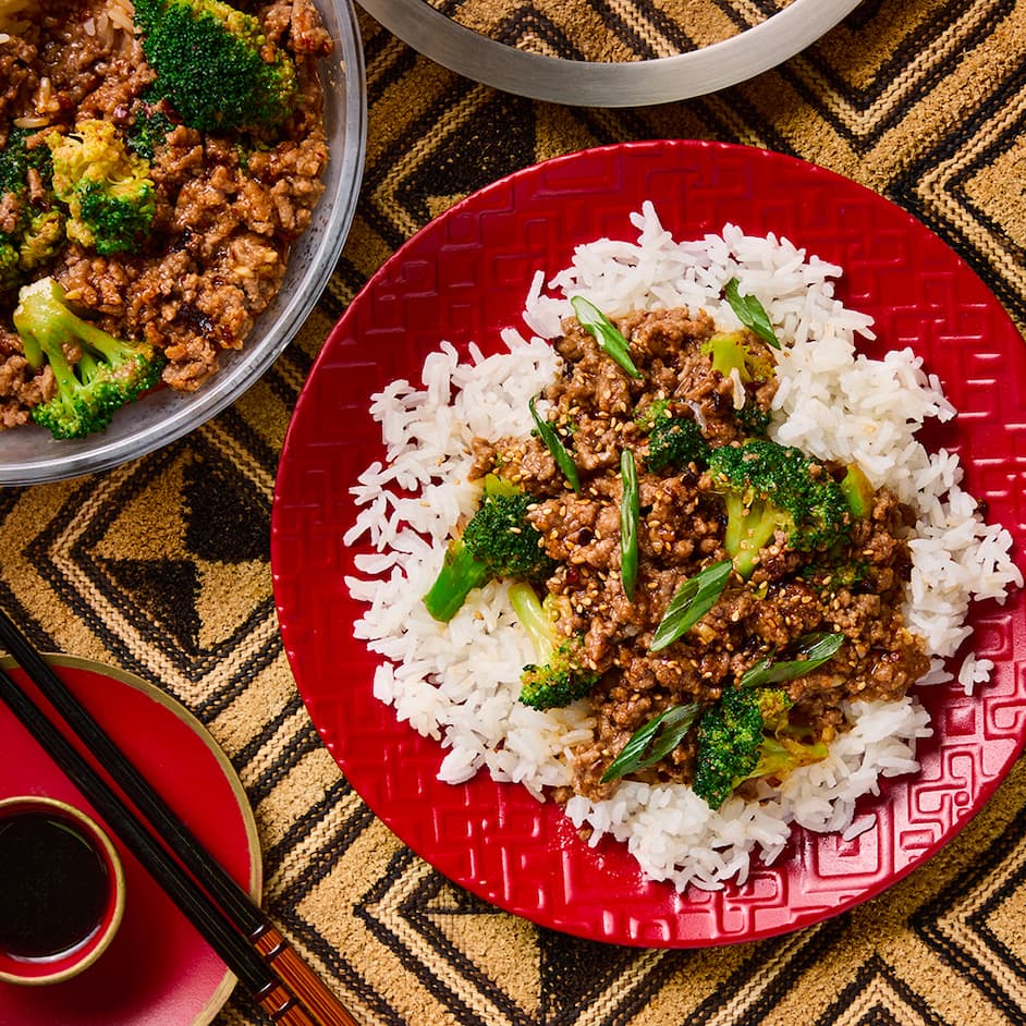 Beef and broccoli served over white rice on a red plate, cooked in the microwave using an Anyday dish. A larger Anyday glass dish with the remaining beef and broccoli is shown nearby on a patterned tablecloth.