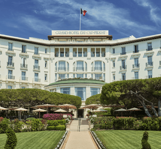 Grand hotel with manicured gardens and a national flag atop, under a bright sky.