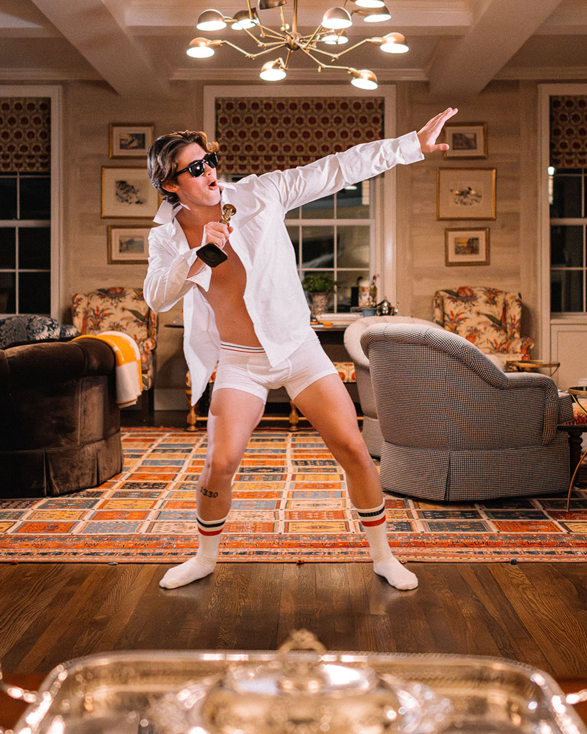 Man wearing white dress shire, white boxers, and white tech varsity socks sliding on hardwood floor holding a trophy as microphone.