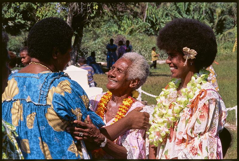 Three people in bright dresses are greeting each other in the sunshine.