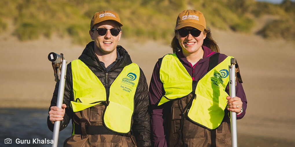 Kyle (left) and Amanda (right) pose in waders and hi-viz yellow vests, at Ocean Beach, ready to test water quality.