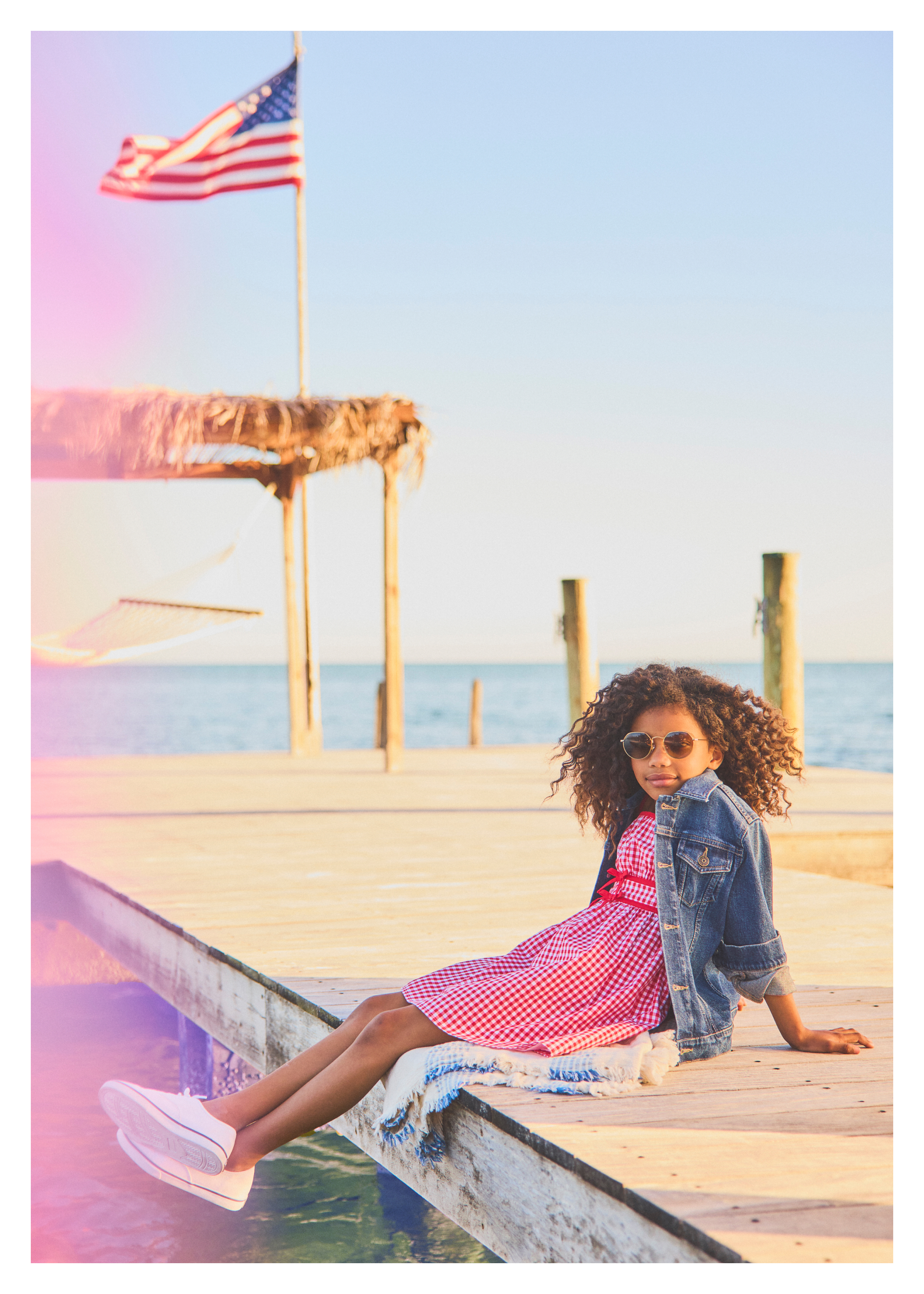 A young girl with curly hair wearing a red gingham dress, denim jacket, and sunglasses sitting on a wooden dock by the ocean with an American flag in the background