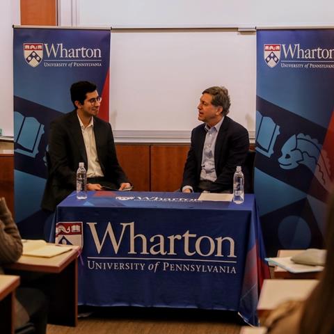 Two speakers in a conversation at a table with Wharton branding.