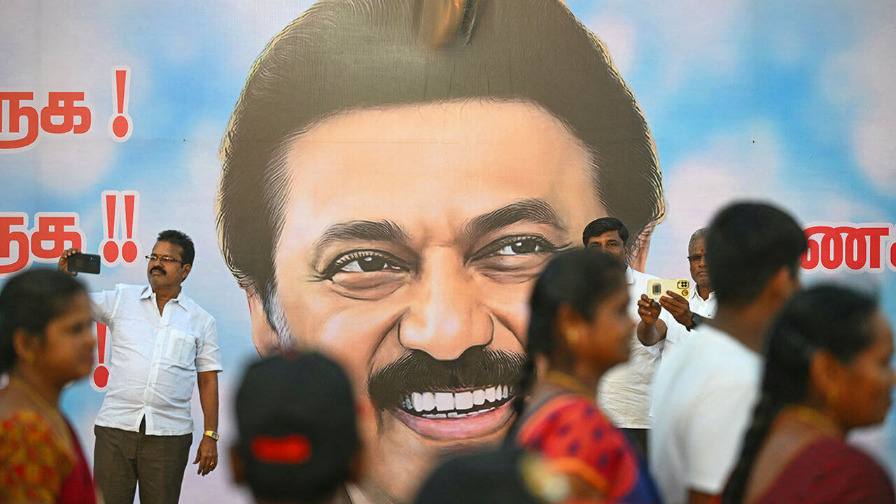 A bilboard of MK Stalin in with background with supporters walking past