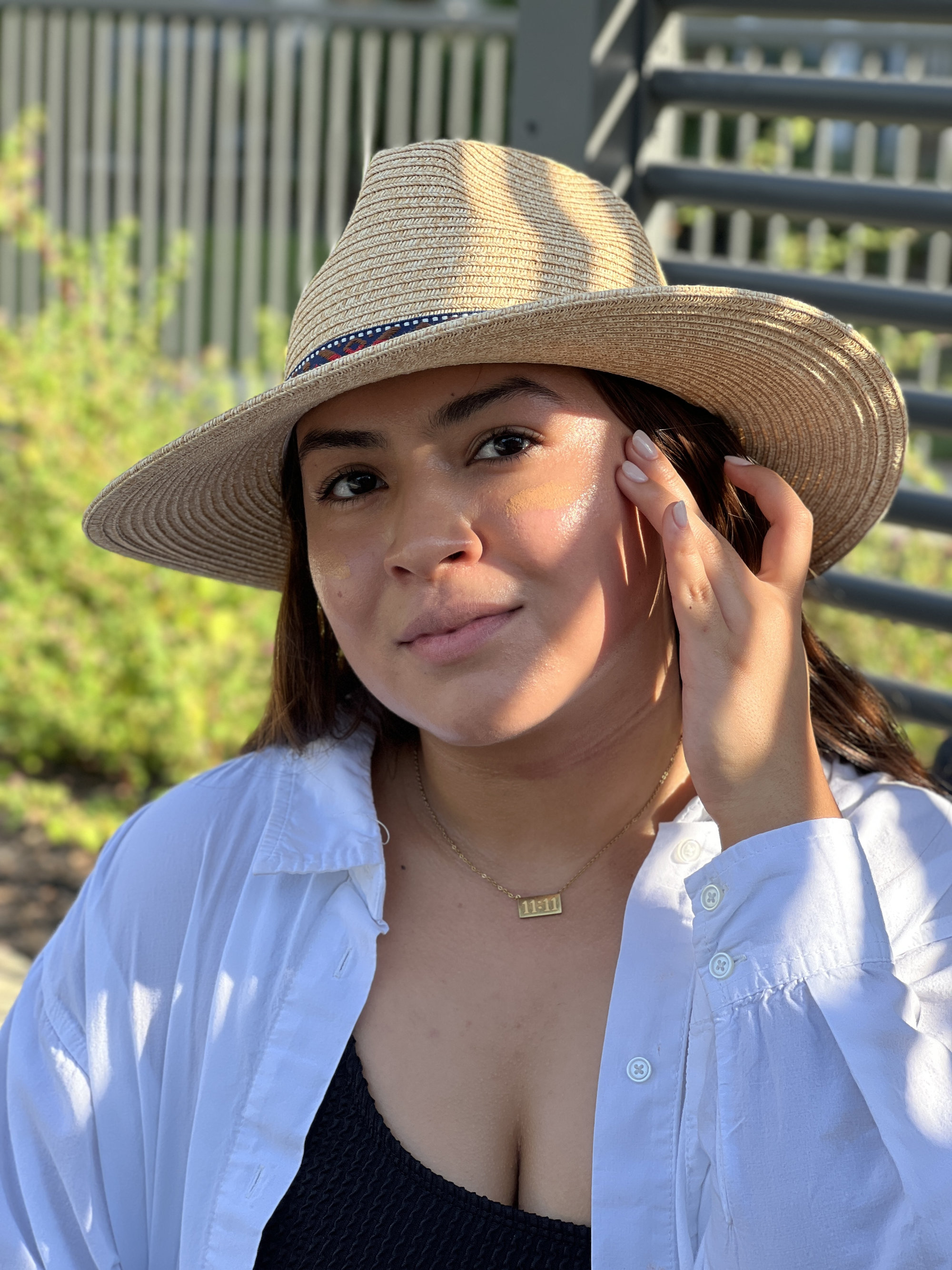 A woman sits in the glow of sunshine wearing a straw hat and a lightweight white shirt.