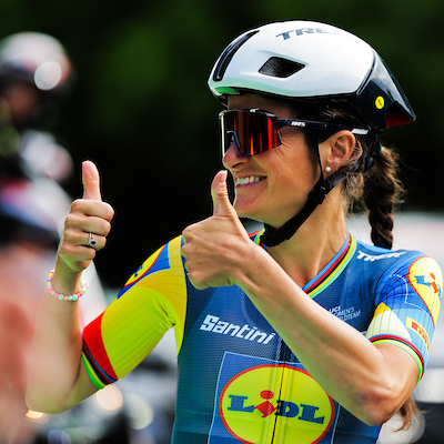 Lizzie Deignan in Lidl Trek team kit, wearing sunglasses and a helmet and giving a double thumbs up to the side.