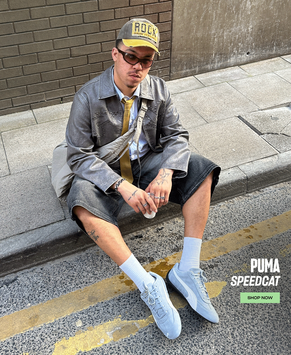 A GIF of two images. The first image shows a person wearing a pair of grey PUMA Speedcat trainers with grey shorts and jacket whilst sitting at a roadside on a street. Second image shows a person holding a mobile phone over their feet whilst wearing a pair of grey PUMA Speedcat trainers. The image contains the text 'Puma Speedcat Shop Now'