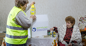 An IFCJ staff member wearing a yellow vest, opening an IFCJ food box with an elderly woman.