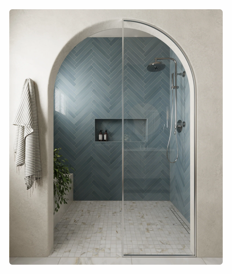 A modern walk-in shower with a curved glass door, featuring light blue subway tiles in a herringbone pattern and a white marble mosaic floor.