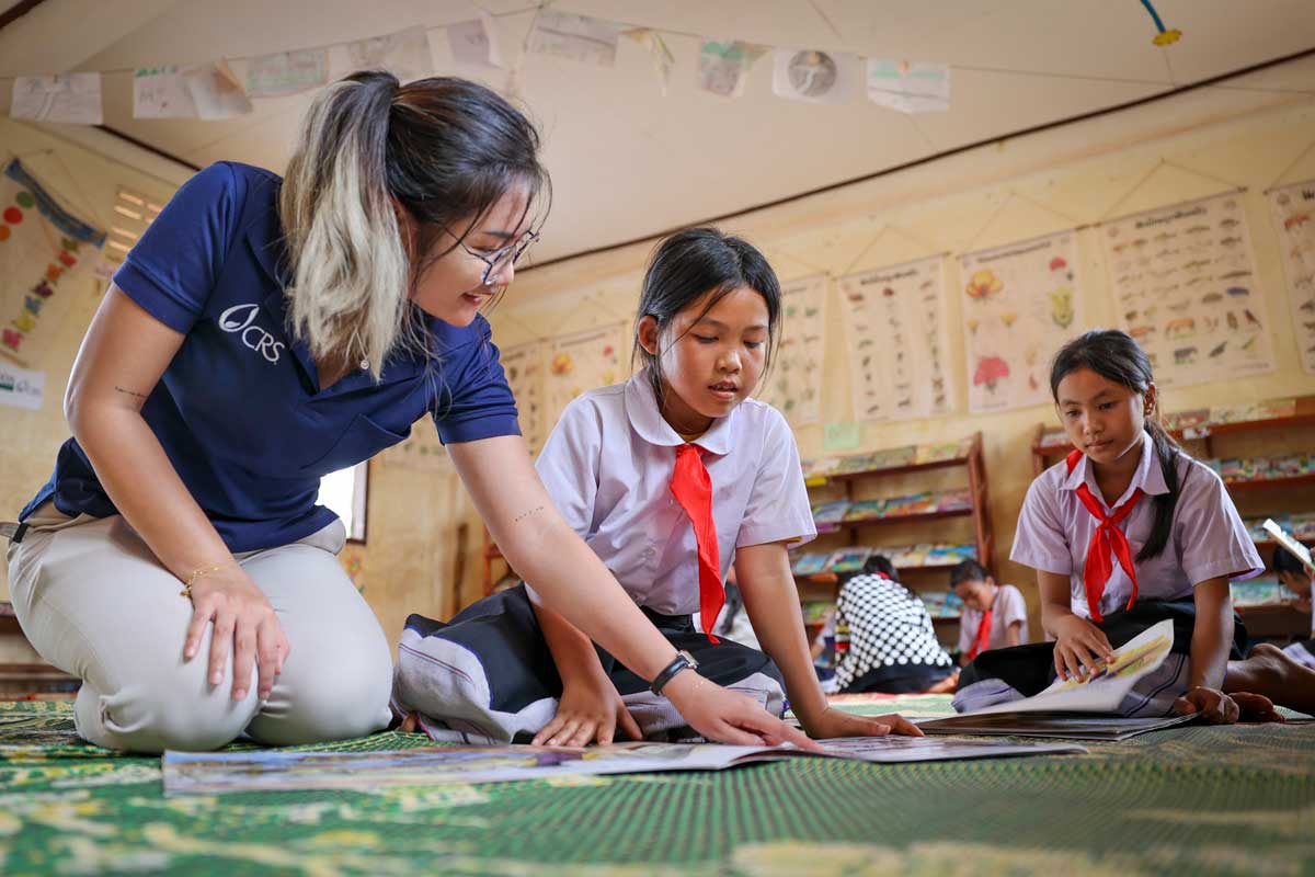 A teacher reads alongside two schoolgirls in a classroom decorated with posters and drawings.