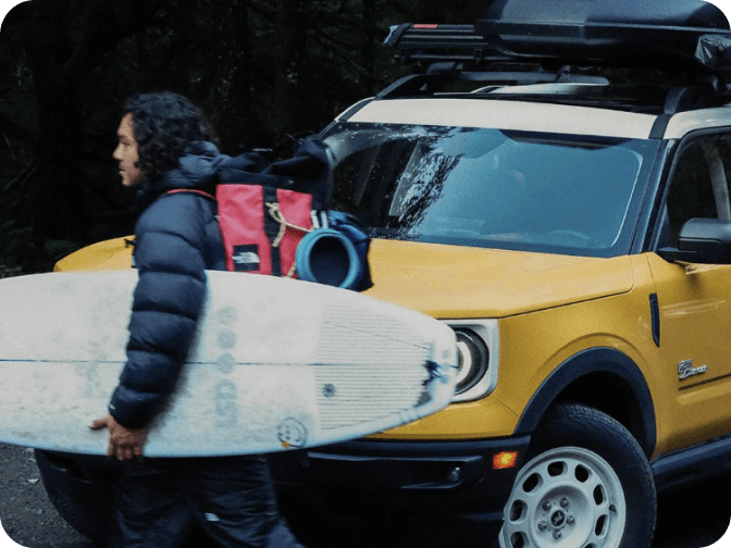 A man wearing a backpack and carrying a surfboard in front of a parked Bronco&reg; Sport SUV