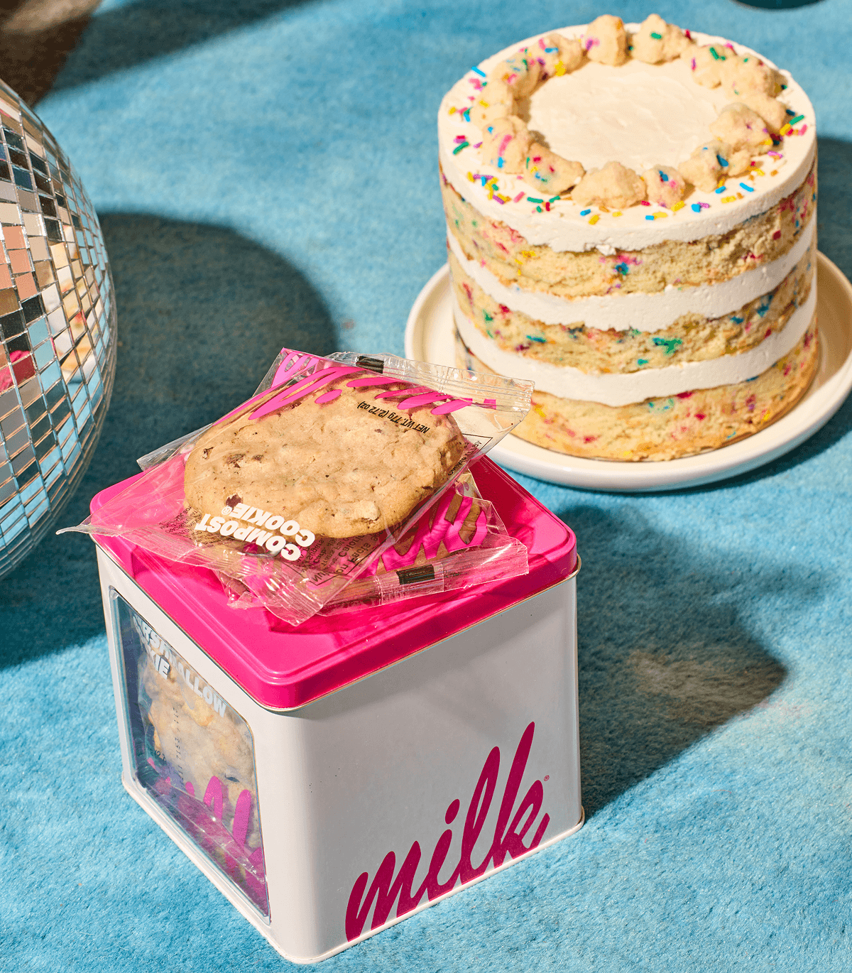 Cookie tin with wrapped cookies in foreground and confetti cake in background next to a disco ball on a blue surface.