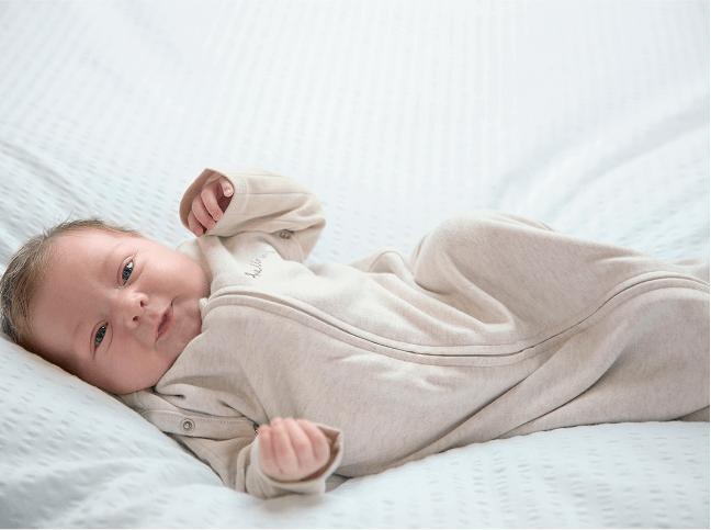 Baby lay, smiling, wearing the oatmeal style swaddle with the arms free mode