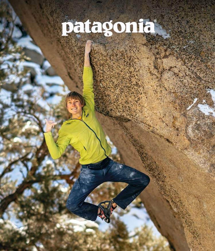 A climber smiles at the camera while hanging from a hold with one hand.