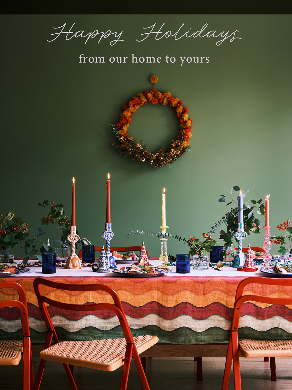 A richly decorated table with bright candles, patterned linens, and greenery sits beneath a wreath on a deep green wall. The warm colours and soft glow evoke a cosy, celebratory atmosphere perfect for gathering and connection.