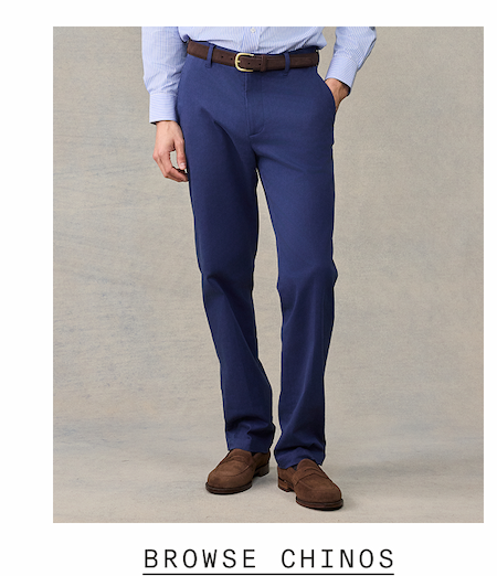Close-up of a man from the waist down wearing slim-fit navy chinos with a brown leather belt and brown suede loafers, paired with a blue striped dress shirt. Text reads: "BROWSE CHINOS"